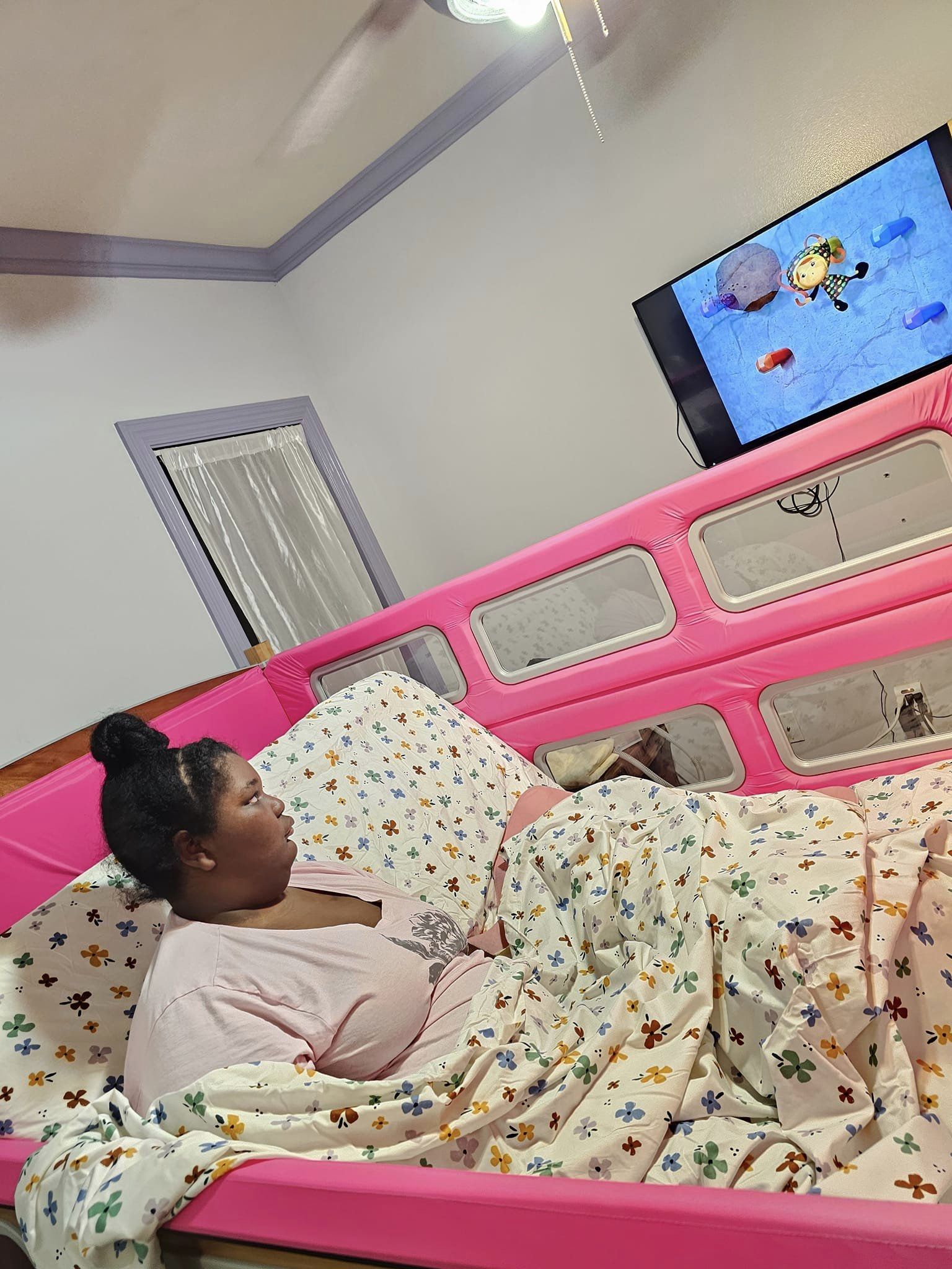 Amoree sitting up in her sleep safe bed with hot pink bedrails watching Team  Umizoomi on the tv on the wall. The walls are light purple with a deep purple crown molding.